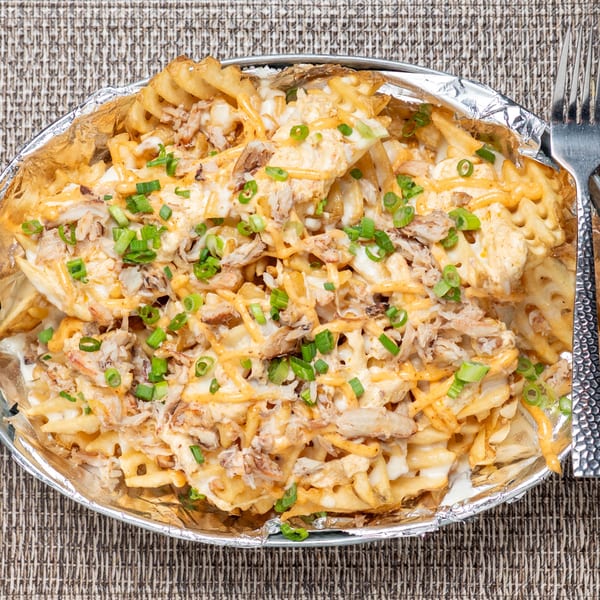 Waffle fries topped with shredded chicken, melted cheese, and green onions in a foil container.
