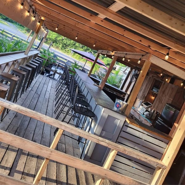 An empty outdoor bar and dining area with wooden floors, string lights, and a view of a road.