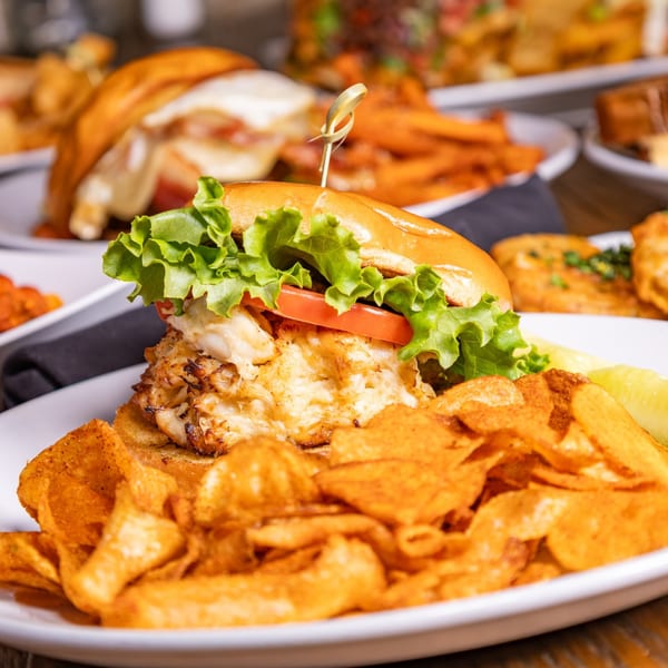 A crab cake sandwich with lettuce and tomato, served with potato chips and a pickle spear.