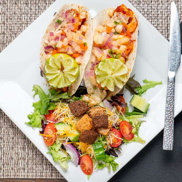 Two shrimp tacos with pineapple salsa and lime wedges, served with a side salad.