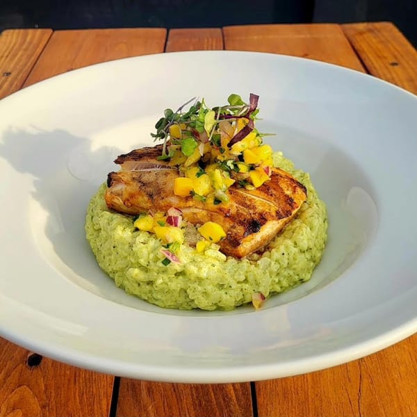 Grilled fish with mango salsa and green risotto, served on a white plate on a wooden table.