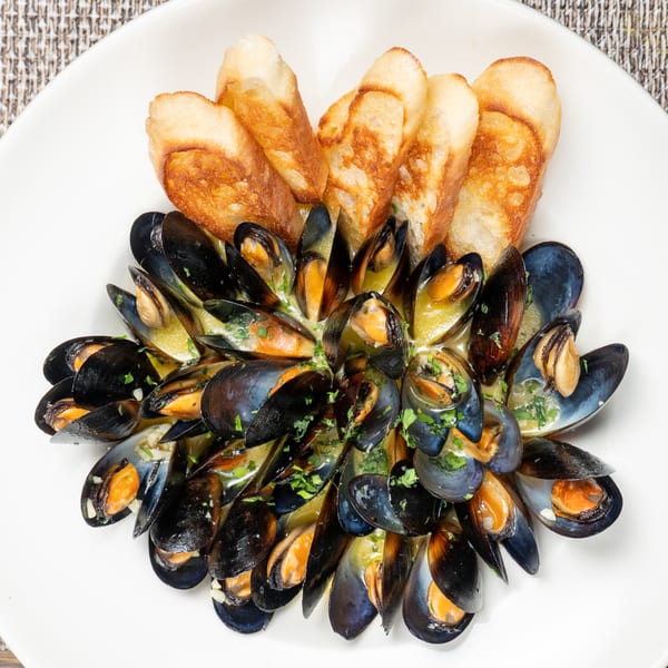 Steamed mussels in a creamy sauce, garnished with parsley and served with toasted bread on a white plate.