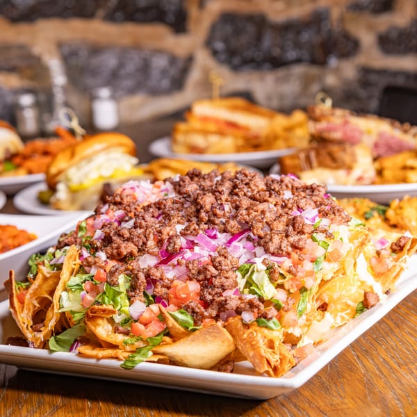 Loaded nachos with ground beef, lettuce, diced tomatoes, and red onions, with other American dishes in the background.
