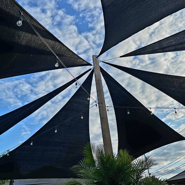 Black shade sails and string lights create a geometric pattern against a blue, cloudy sky.