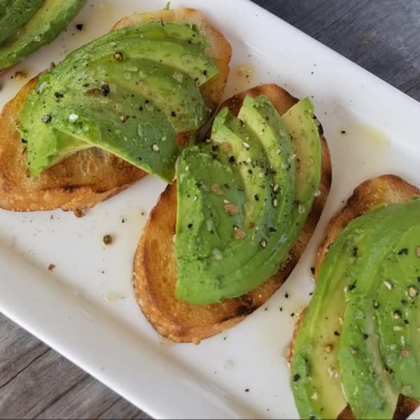 Avocado toast with sliced avocado, black pepper, and olive oil on a white rectangular plate.