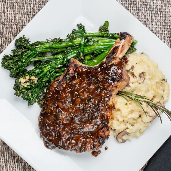 Grilled pork chop with dark sauce, broccolini, and mushroom risotto on a white plate.