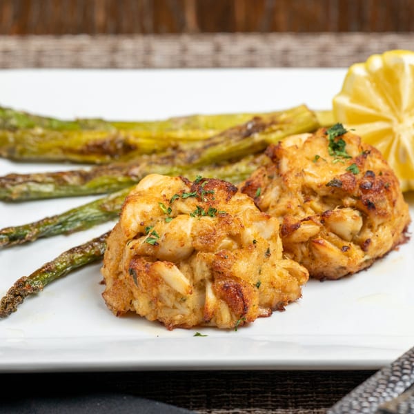 Two golden-brown crab cakes with roasted asparagus and a lemon wedge on a white plate.
