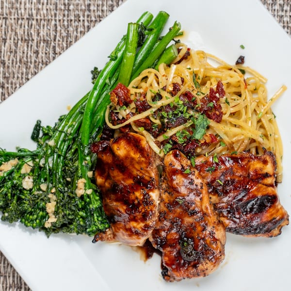Grilled chicken with broccolini and pasta topped with sun-dried tomatoes and herbs on a white plate.